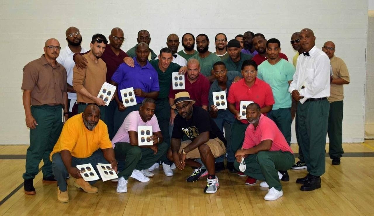 Freedom Reads Founder & CEO Reginald Dwayne Betts, front row in a fedora, with audience members at Otisville Correctional Facility in New York holding copies of Dwayne's poetry collection FELON.