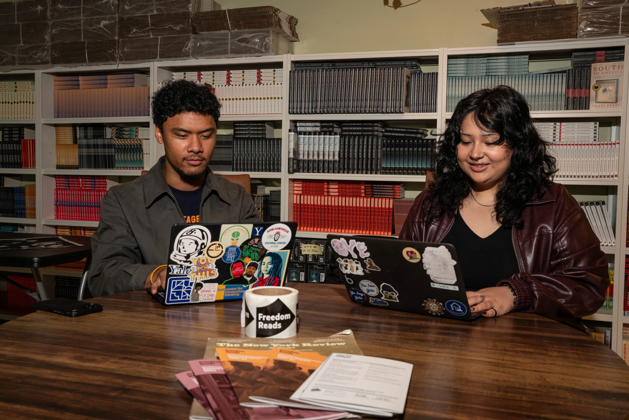 Young man and a woman looking at laptops in a room with bookshelves.
