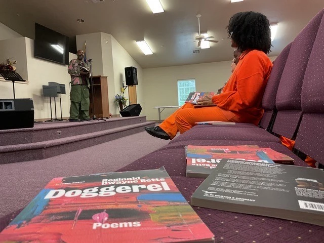 Doggerel books lay on auditorium chairs in a prison, women in the background in orange prison uniforms sitting watching Dwayne Betts perform on a small stage.