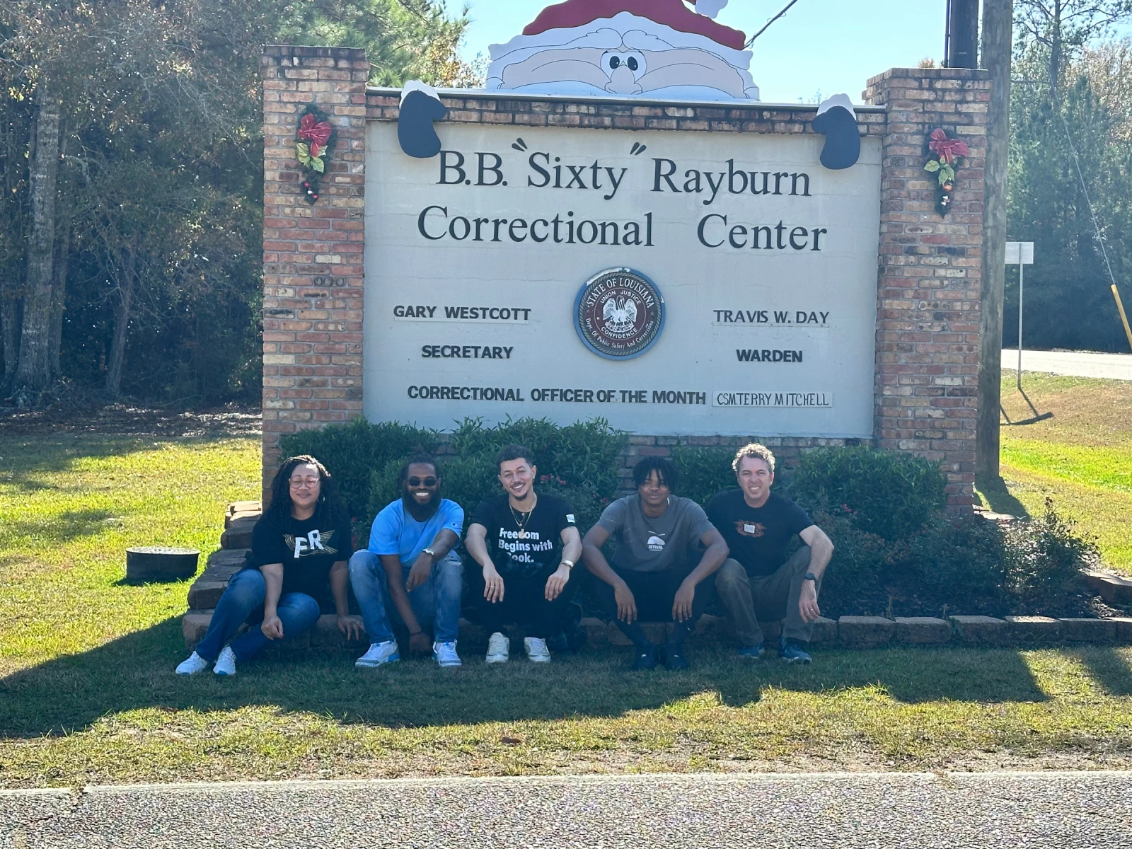 Five members of the Freedom Reads team sit in front of the sign for Rayburn Correctional Center wearing Freedom Reads t-shirts.