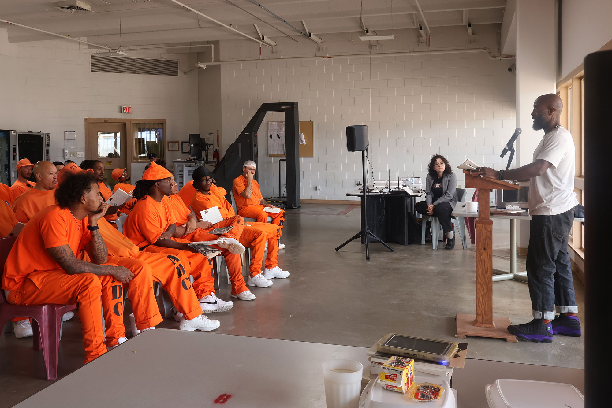 A group of Arizona State Prison Yuma inmates clad in bright orange listening to Dwayne Betts speaking at a wooden podium.