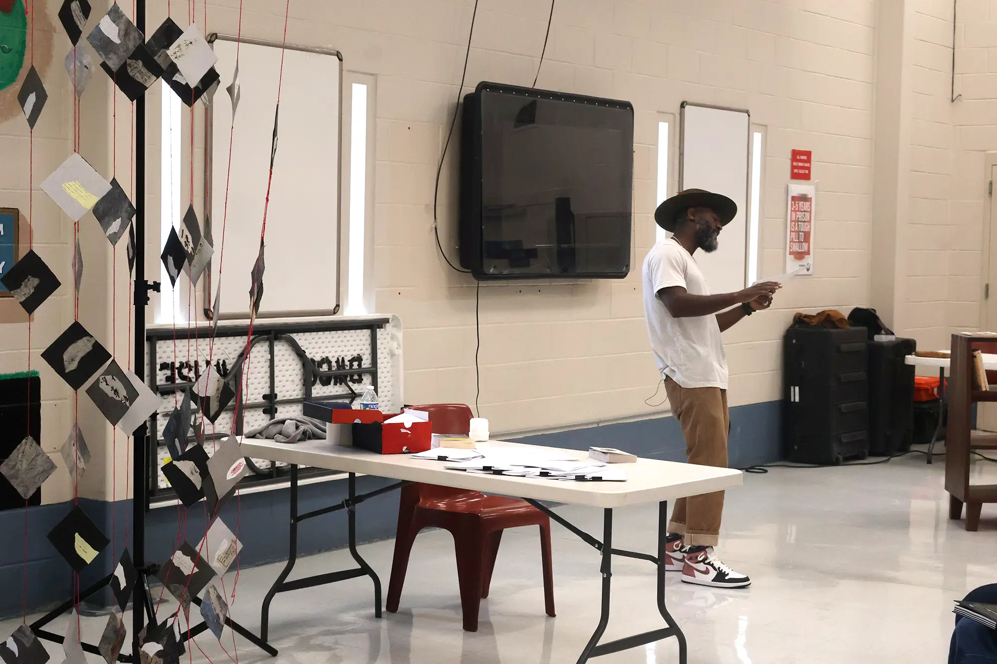 Reginald Dwayne Betts performs among institutional table and chairs between hanging works made from traditional Japanese washi paper as well as a wooden Freedom Library bookcase at Dillwyn Correctional Center in Virginia.