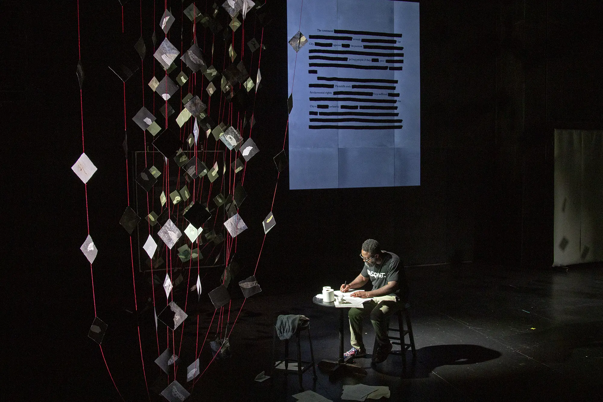 Reginald Dwayne Betts performs, spotlit sitting at a table within an dark installation of visual projection and “kites”, letters and materials made of Japanese washi 