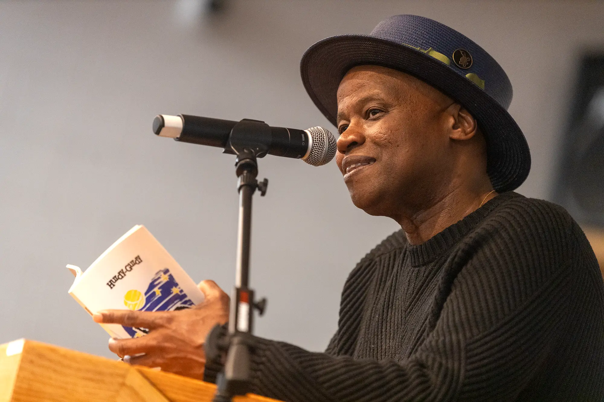Poet Randall Horton wearing a navy boater hat and black sweater, smiles by a microphone at a podium, holding a copy of Hurdy Gurdy by Tim Seibles.